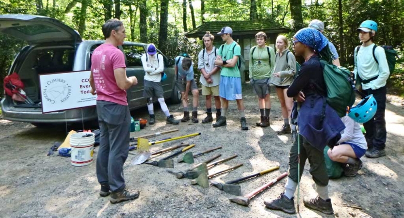 A person speaks to a group of students. There are garden tools laying on the ground between them.